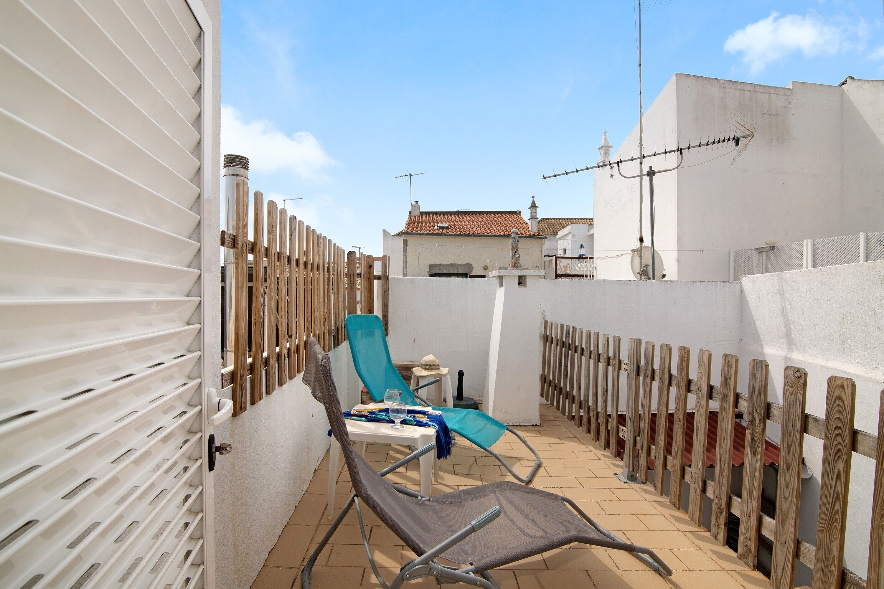 A rooftop terrace with a white wall, a wooden fence, a blue chair, a table with a glass, and a tiled floor.