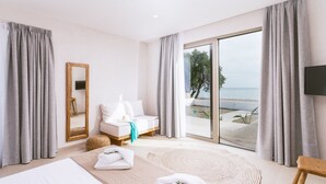 Une chambre d’hôtel moderne dotée d’un grand lit, d’un coin salon avec un canapé et un fauteuil, d’une petite table, d’un miroir et offrant une vue sur la mer grâce à de grandes fenêtres.