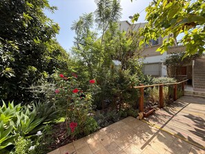 Un jardín con terraza de madera, plantas diversas y un perro pequeño.
