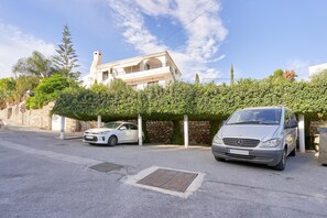 A residential street with two parked cars, a stone wall, and a building with a white facade and arched windows.