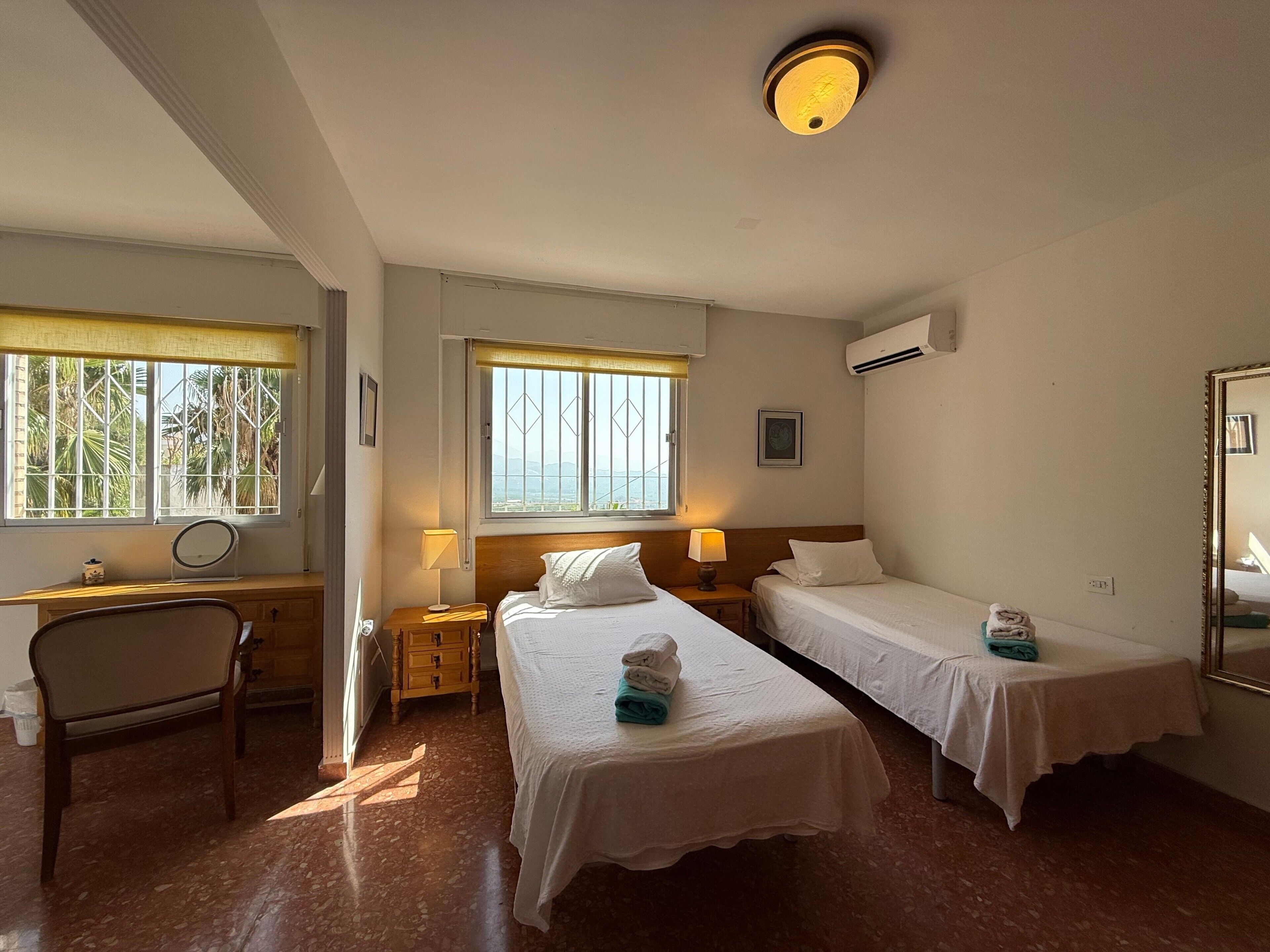 A hotel room with two single beds, a desk with a chair, a window with a view, and an air conditioning unit.