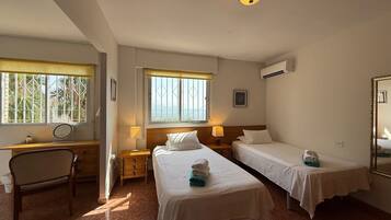 A hotel room with two single beds, a desk with a chair, a window with a view, and an air conditioning unit.