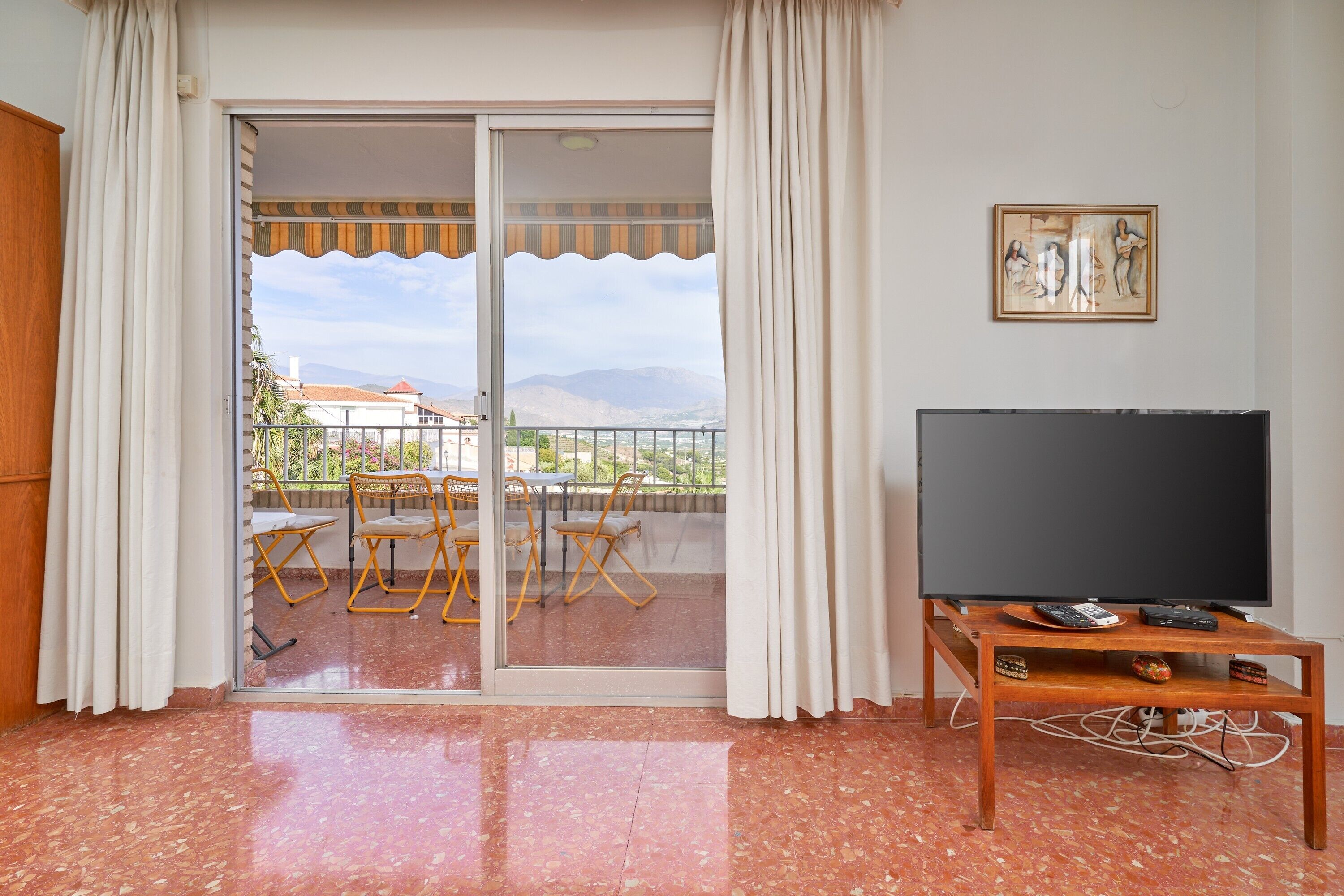A room with a television on a wooden stand, a sliding glass door leading to an outdoor area with chairs and a table, and a painting on the wall.