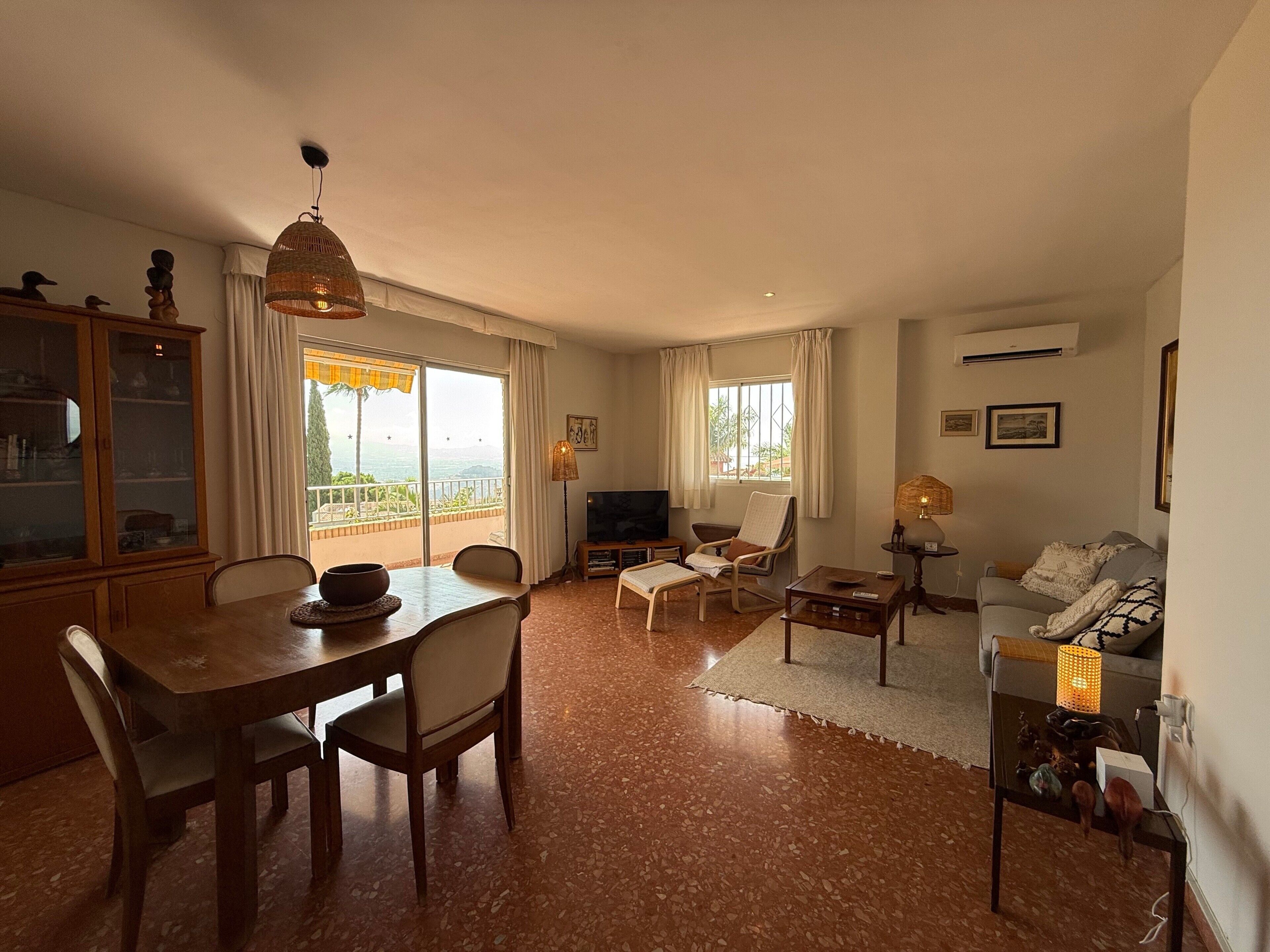 A living room with a dining area, a TV, and a balcony with a view.