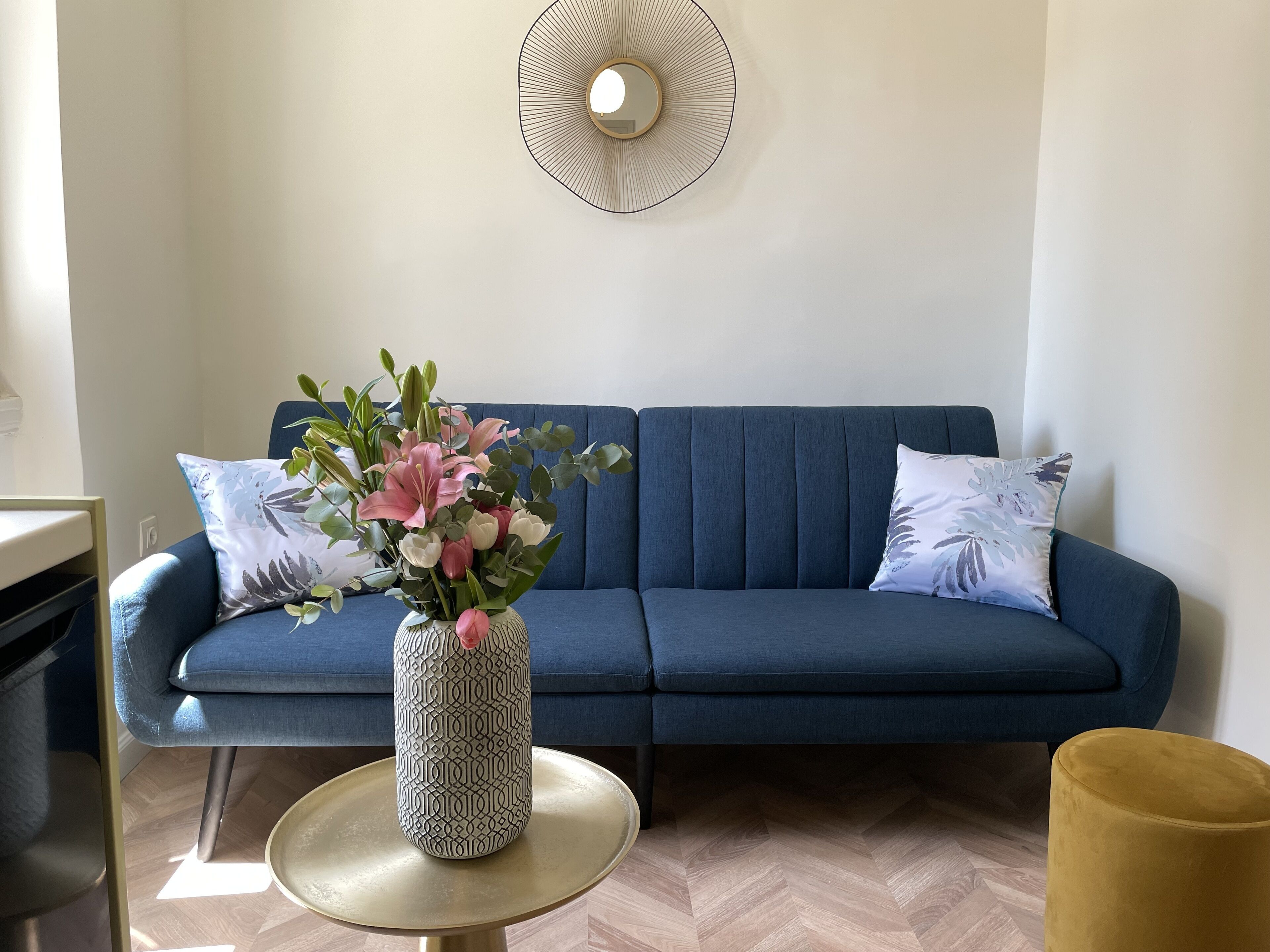 A modern living room with a blue sofa, a patterned vase with flowers, and a round gold table.