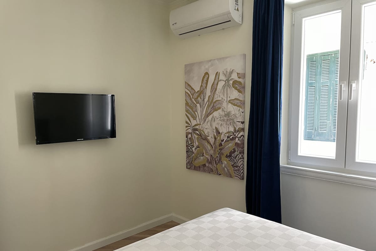 A bedroom with a bed, a mounted flat-screen TV, a wall-mounted air conditioner, a window with blue curtains, and a floral artwork on the wall.