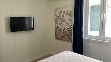 A bedroom with a bed, a mounted flat-screen TV, a wall-mounted air conditioner, a window with blue curtains, and a floral artwork on the wall.