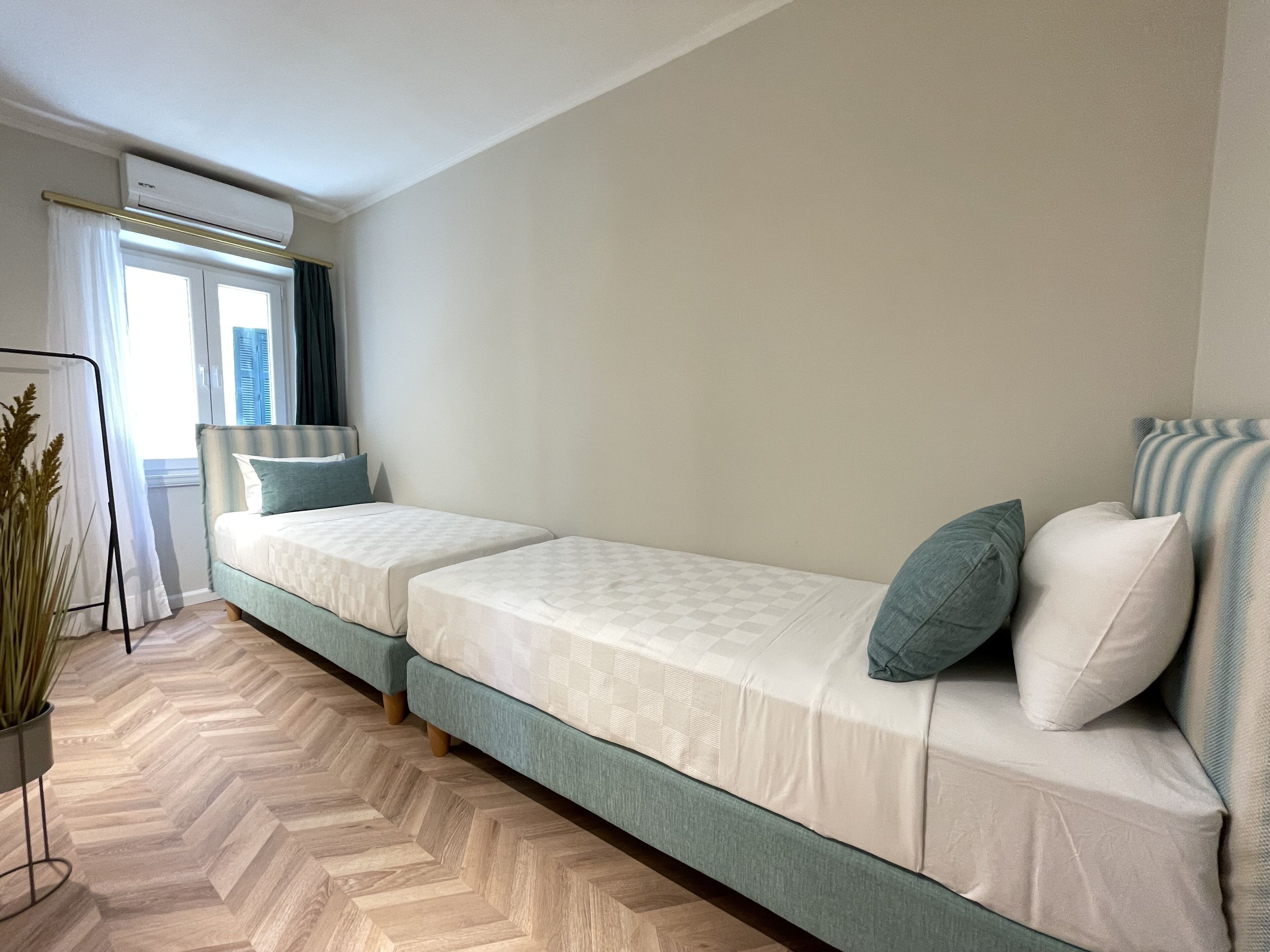 A room with two single beds, a standing heater, a window with curtains, and a wooden herringbone floor.
