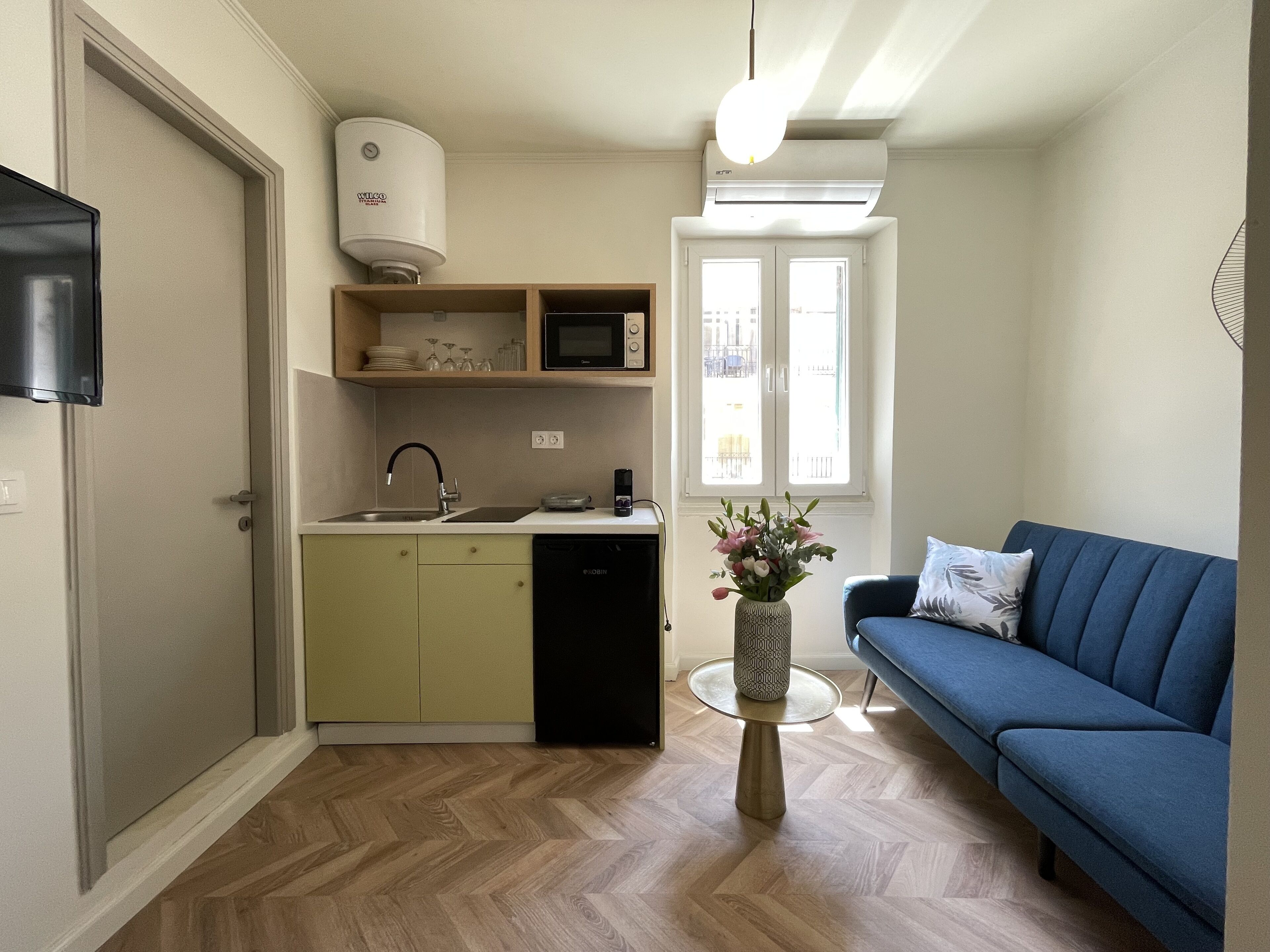 Un espace de vie compact comprenant un coin cuisine, un canapé bleu et une petite table ronde agrémentée d’un vase.