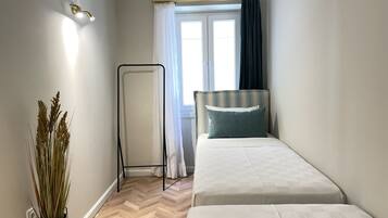 A small, well-lit room with a bed, a bedside table, a potted plant, a window with curtains, and an air conditioning unit.