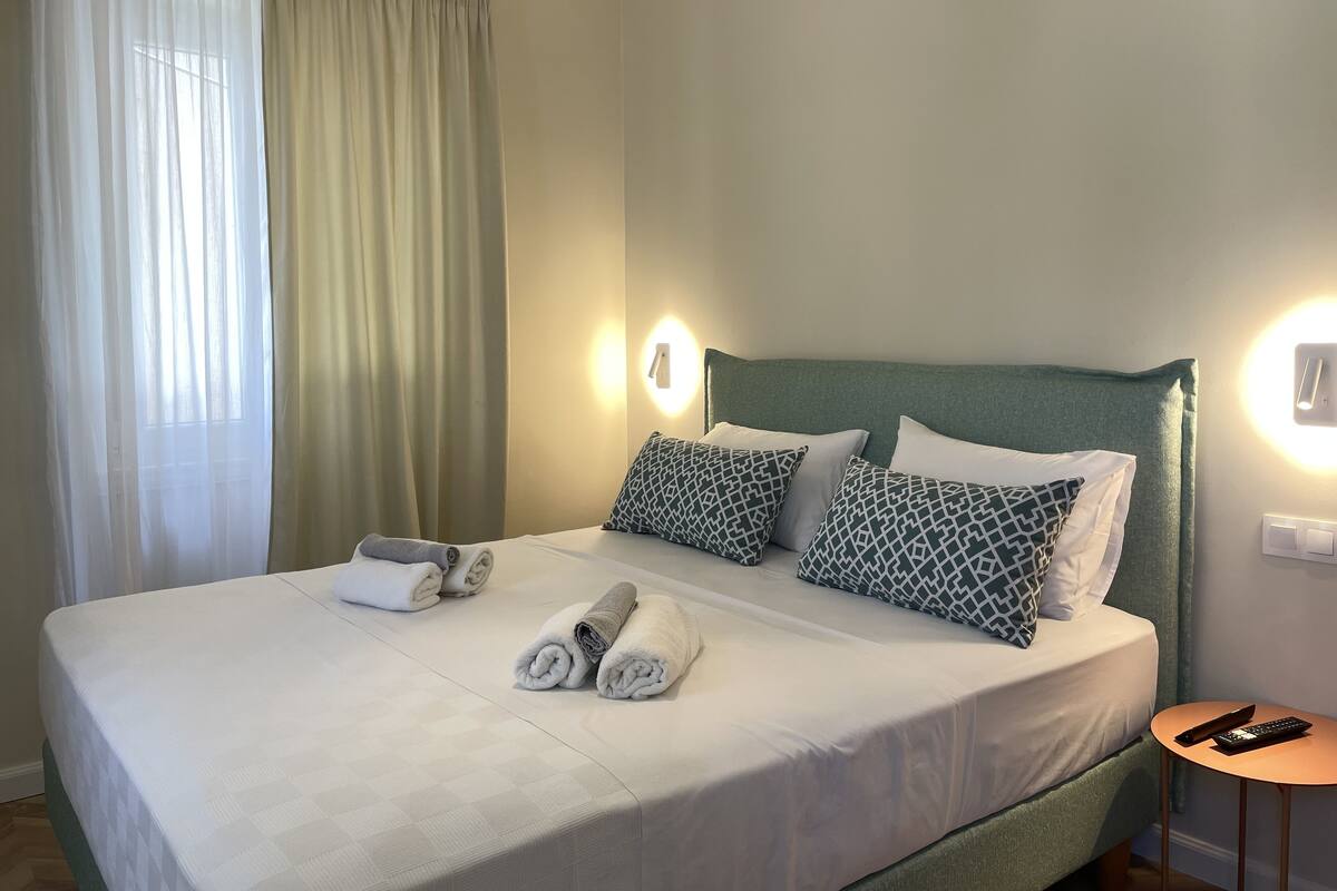 A neatly made bed with rolled towels, a green headboard, patterned pillows, and a remote on a small table beside the bed.