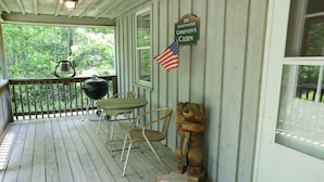 A porch with a wooden deck, a grill, a chair, a bear sculpture, and a cabin sign.