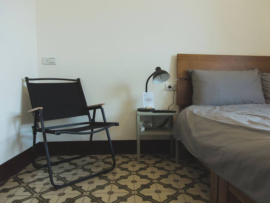 A small, neatly arranged room with a bed, a chair, a desk with a lamp, and a patterned floor.