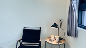 Een kleine kamer met een patroon vloer, een enkele stoel, een tafel met een lamp, en een raam met gordijnen.