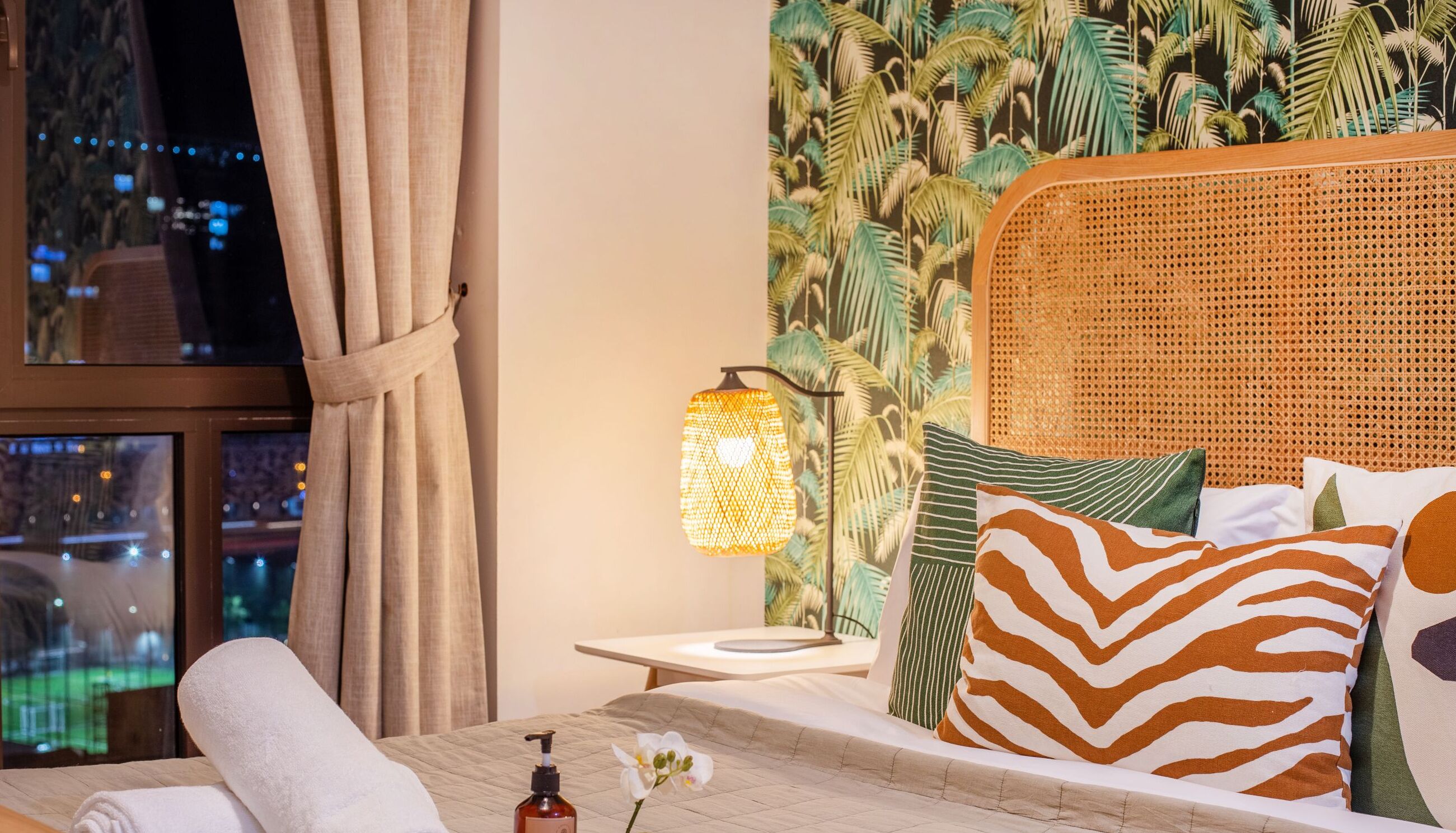 A hotel room with a bed, a nightstand, a lamp, and a view of the city at night.