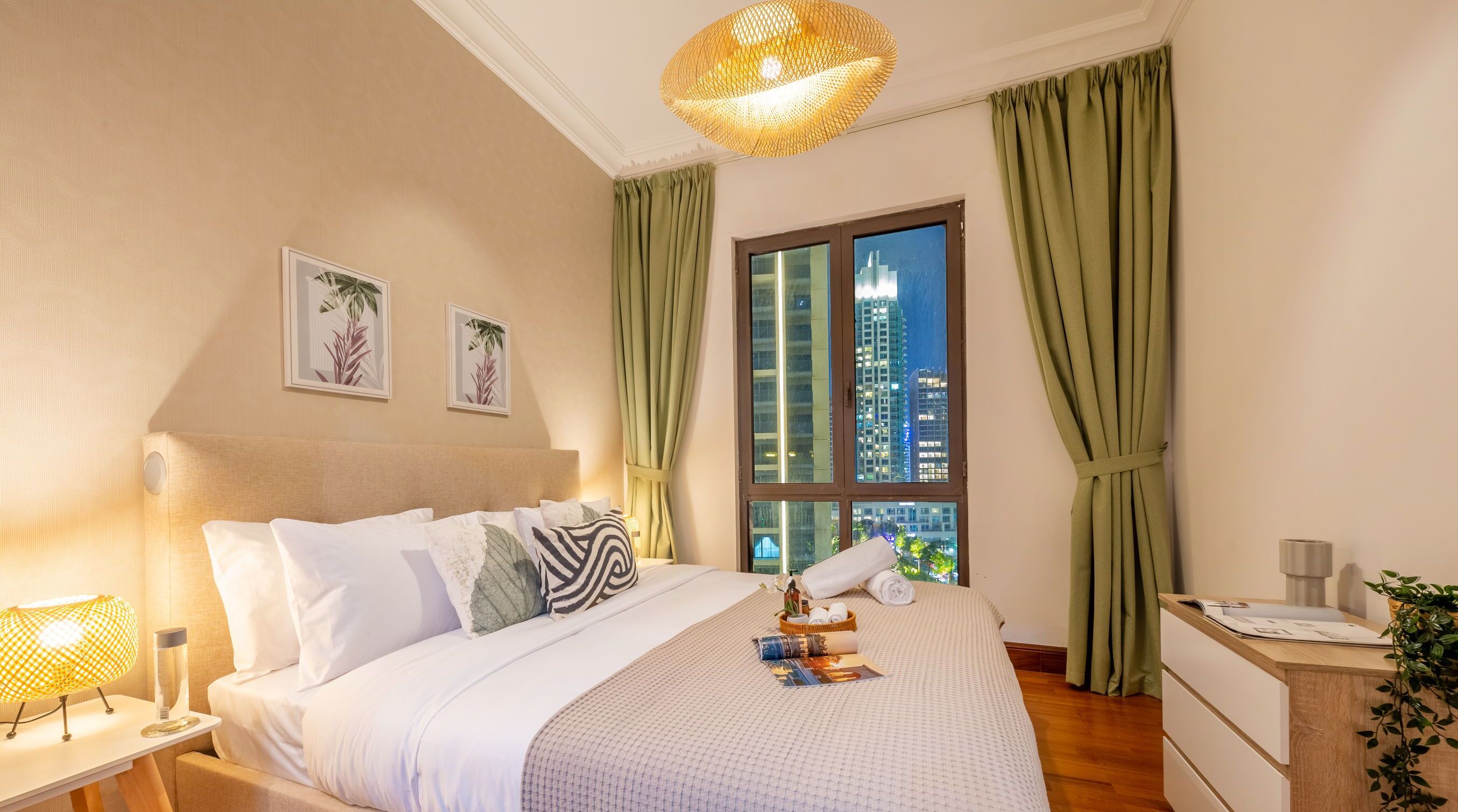 A hotel room with a large bed, a nightstand, a lamp, a window with curtains, and a view of a cityscape.