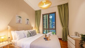 Ein Hotelzimmer mit einem großen Bett, einem Nachttisch, einer Lampe, einem Fenster mit Vorhängen und Blick auf eine Stadtlandschaft.