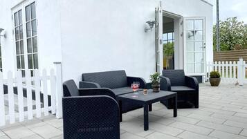 Ein Sitzbereich im Freien mit Korbmöbeln und einem Glastisch, weißen Wänden und einem weißen Zaun.