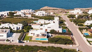 A coastal residential area with modern white buildings, a swimming pool, and a road leading to the sea. - Villa Mercedes - Warm and quiet villa without equal (Ciutadella de Menorca)