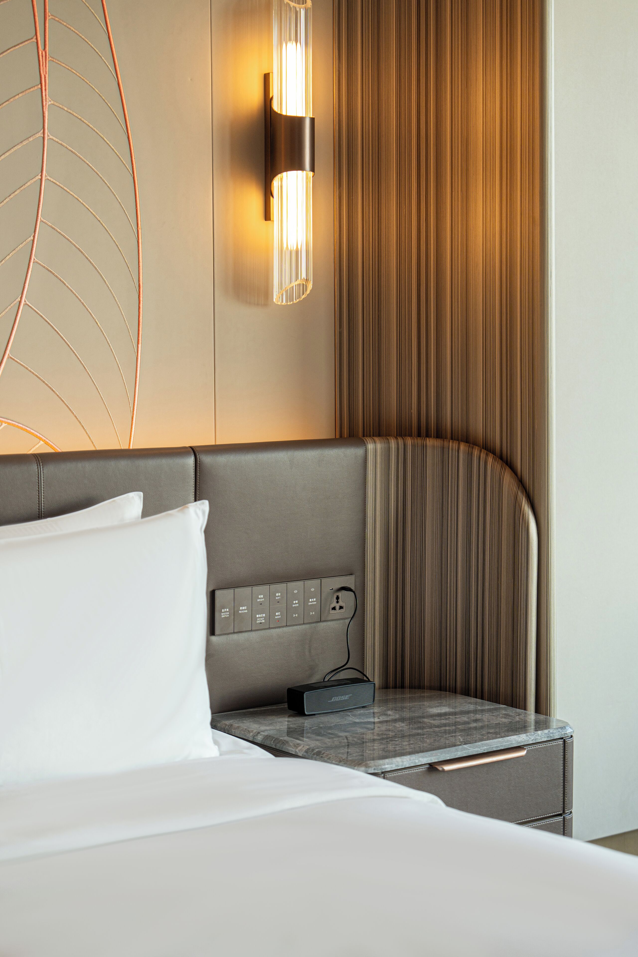 A modern hotel room with a curved headboard, a bedside table with a power outlet, and a wall-mounted light fixture.