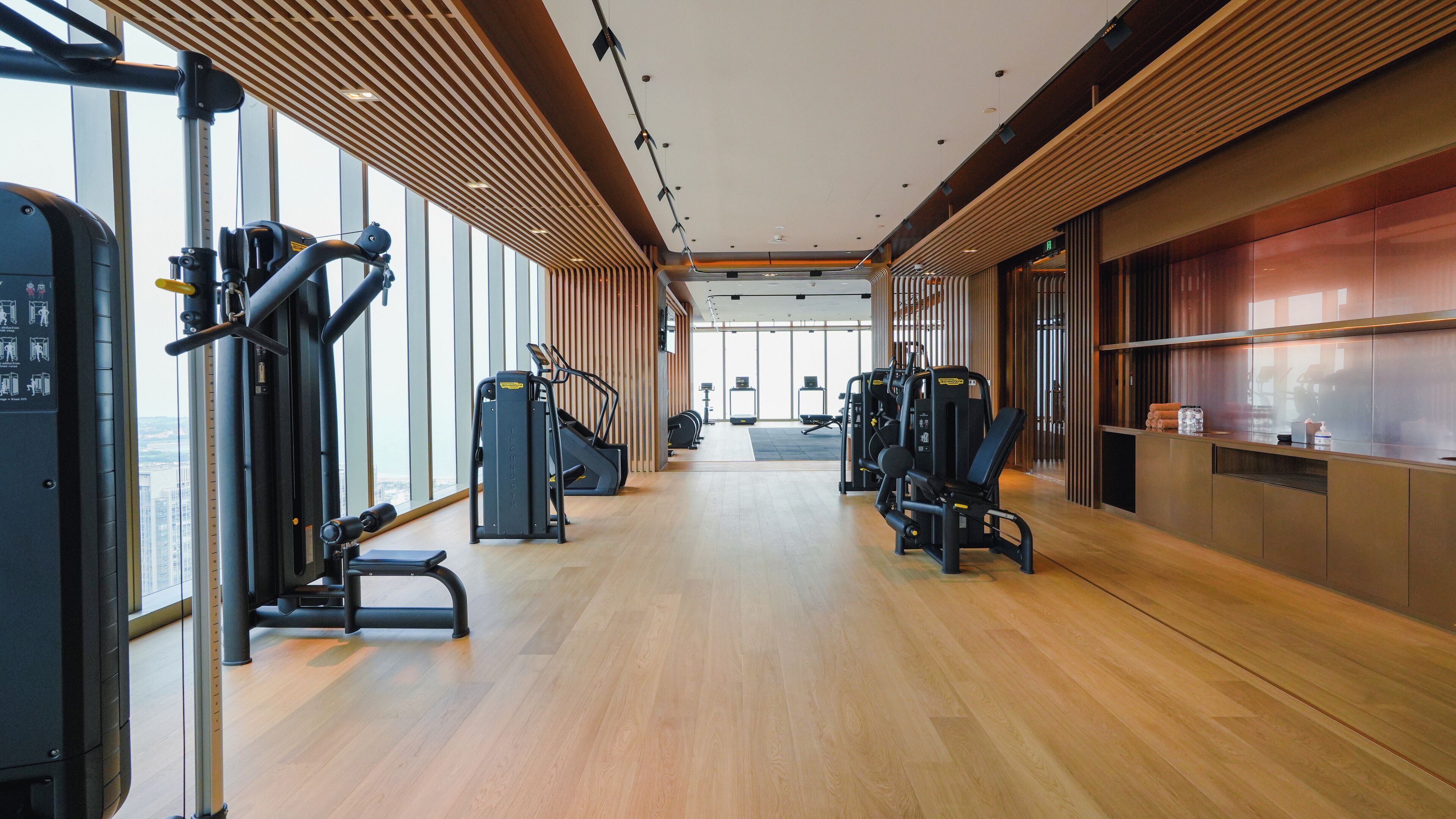 A modern gym with cardio machines, weight stations, and a large window offering a city view.