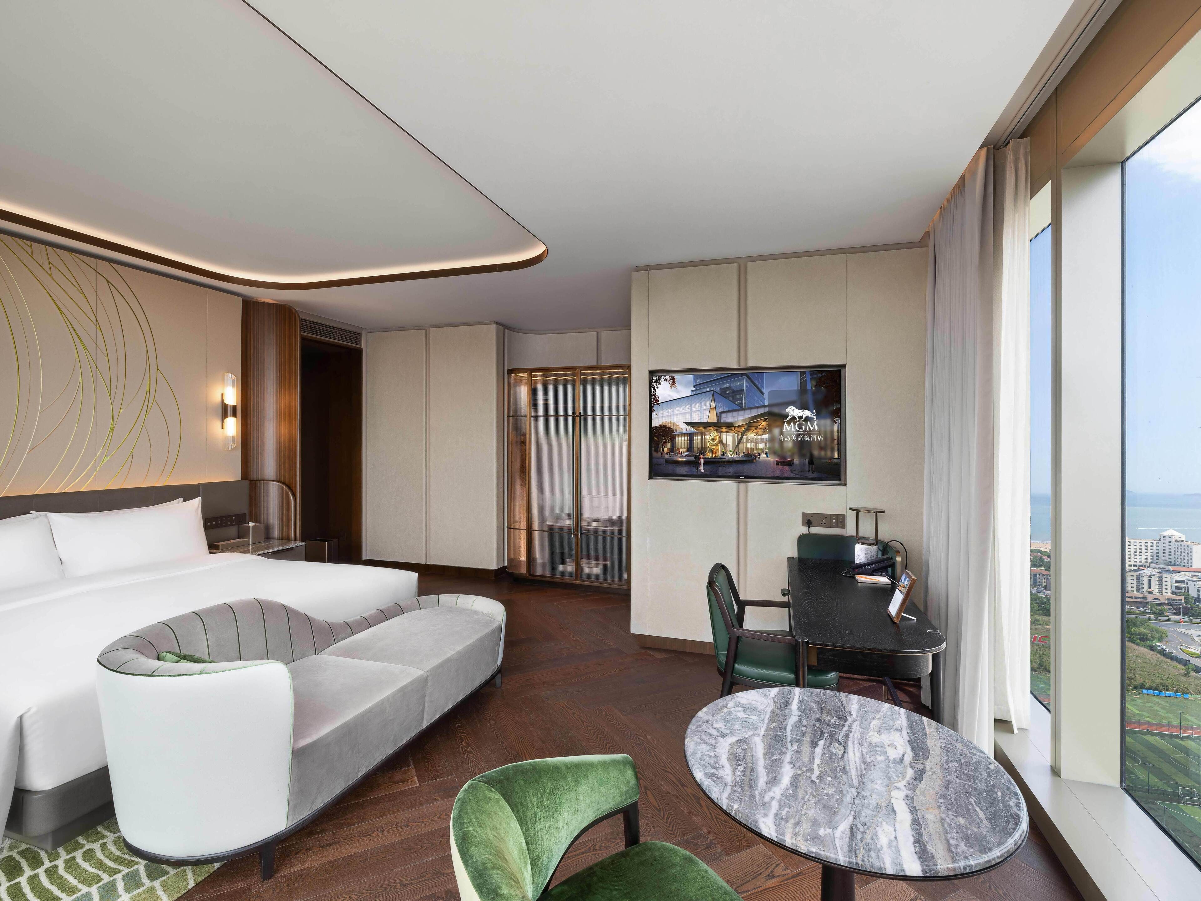 Una habitación de hotel moderna con un sofá curvo, una mesa redonda de mármol, un sillón verde, un televisor montado en la pared y vistas de la ciudad a través de amplias ventanas.