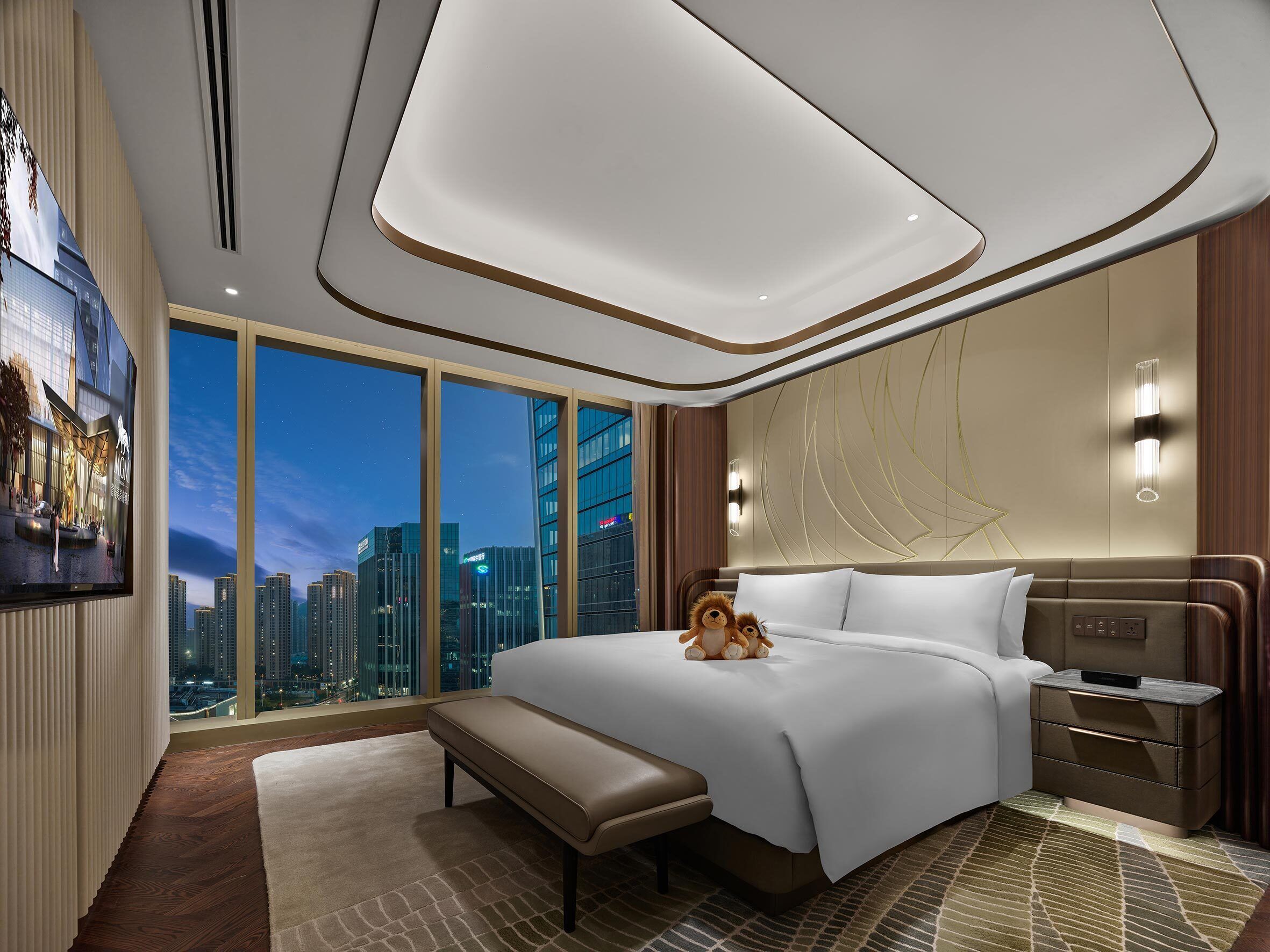 A modern hotel room with a large bed, a bench, a nightstand, and a city view.
