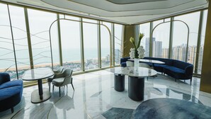 A modern lounge with a panoramic city and beach view, featuring a marble floor, blue sofas, and a glass table. - MGM Qingdao (Qingdao)