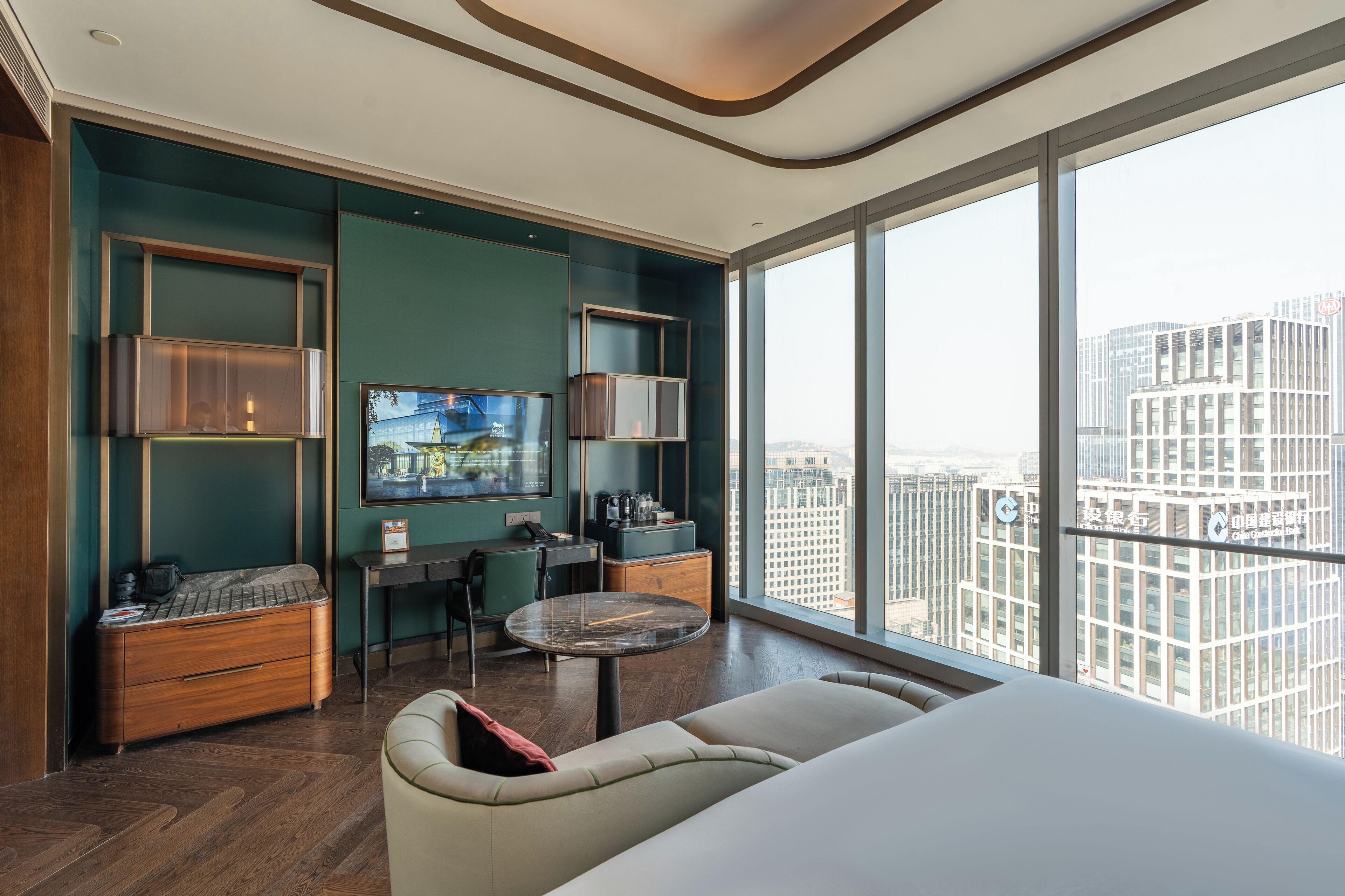 A modern hotel room with a large window, a flat-screen TV, a dining area, and a sofa.
