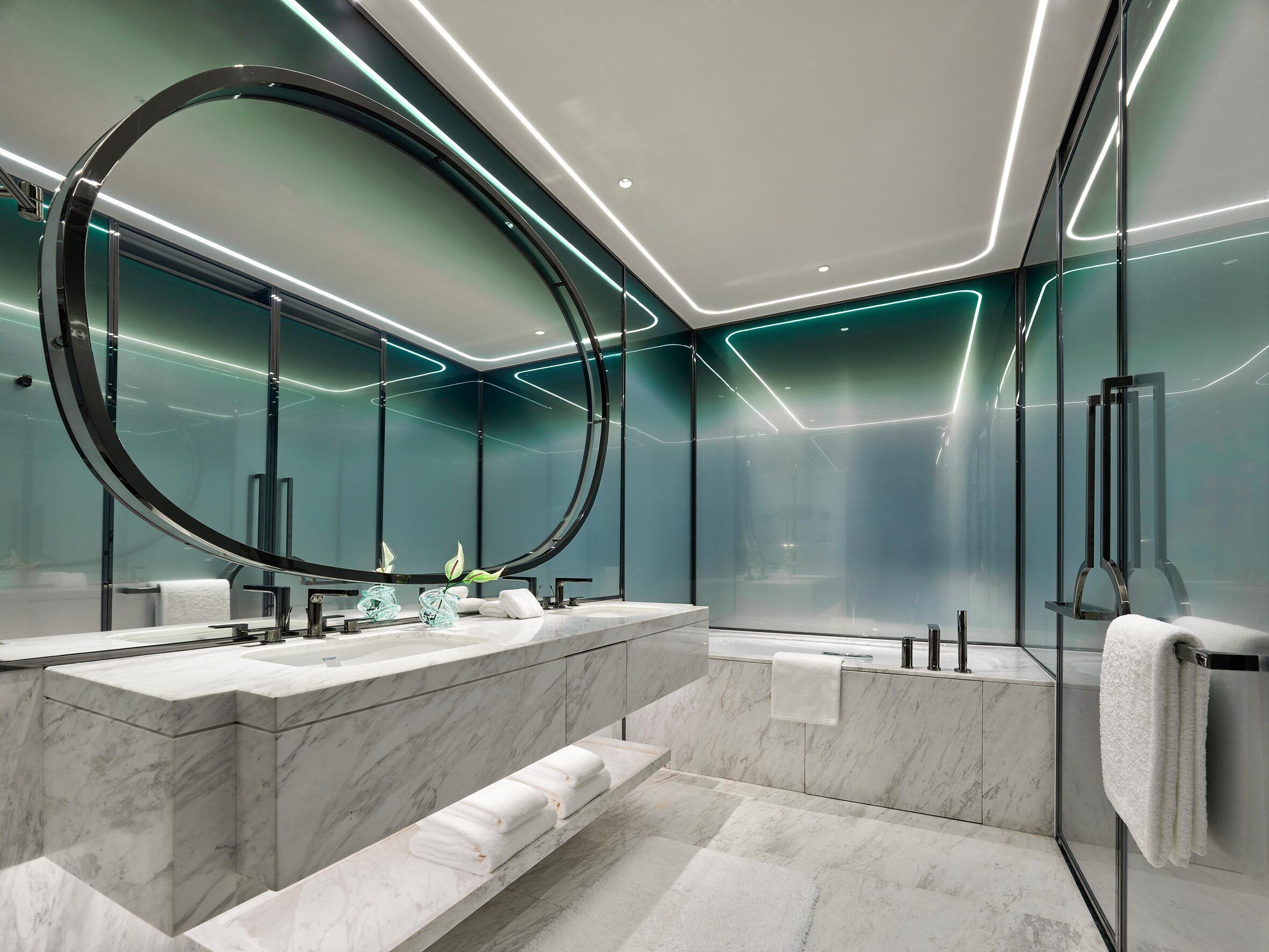 A modern bathroom with a large oval mirror, marble countertops, and a glass-enclosed shower area.