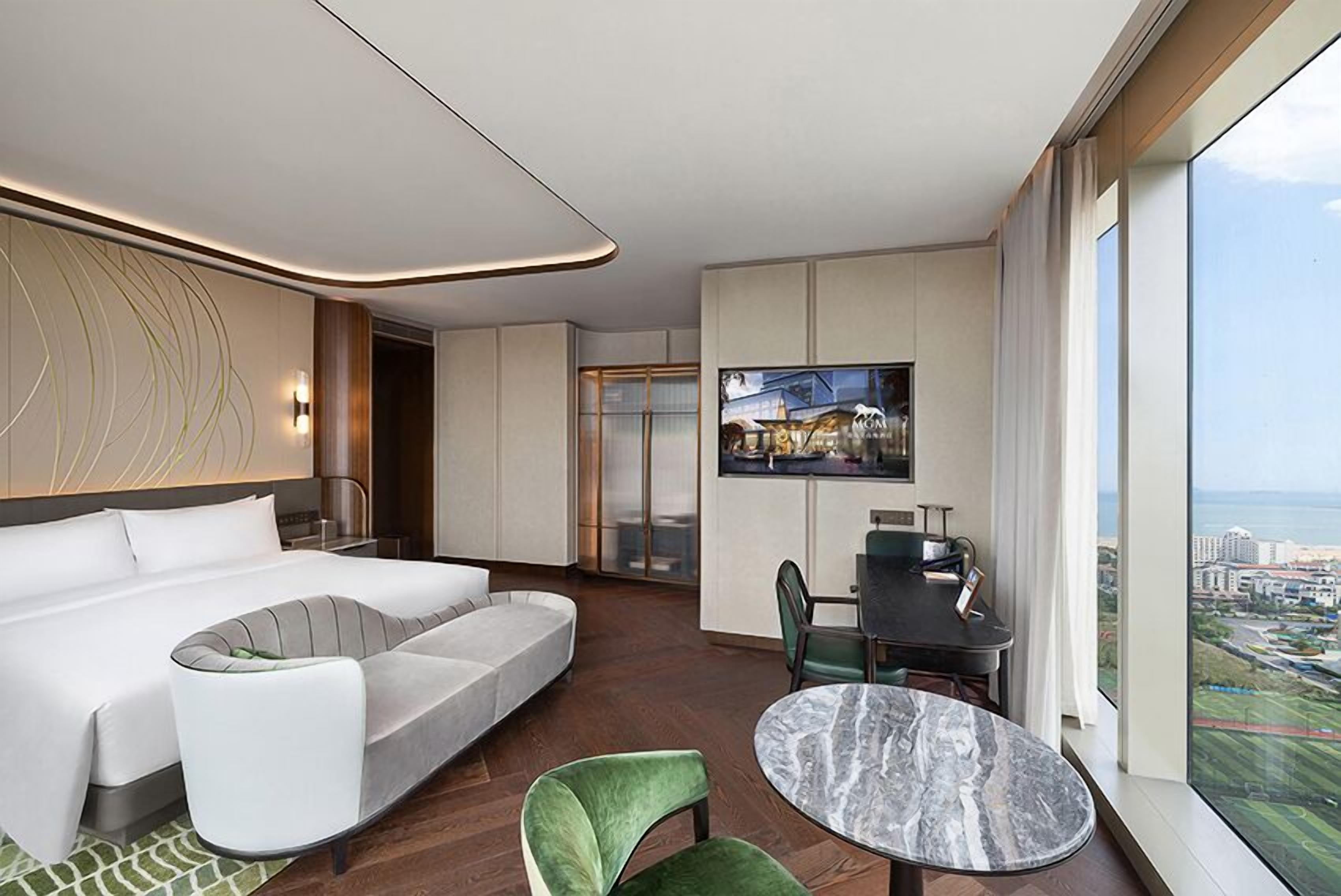 A modern hotel room with a large bed, a sofa, a dining table, and a view of the city.