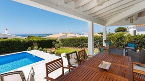 A covered outdoor area with a pool, wooden furniture, and a view of the sea. - Bini Sole - Luxury villa with pool in Menorca (Sant Lluís)