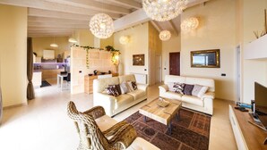 A spacious living room with a sofa set, a coffee table, and wicker chairs. There are pendant lights hanging from the ceiling, a fireplace, and a kitchen area in the background. - Bini Sole - Luxury villa with pool in Menorca (Sant Lluís)