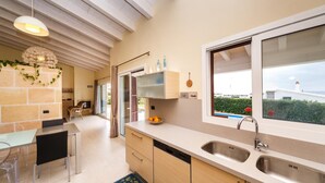 A modern kitchen with a large window, a built-in sink, and a dining area with a glass table and chairs. - Bini Sole - Luxury villa with pool in Menorca (Sant Lluís)
