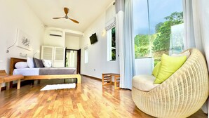 Una habitación con una cama, una silla de mimbre, un ventilador de techo y ventanas amplias con vistas a la vegetación.