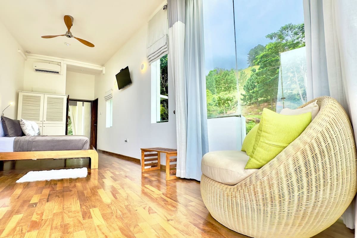Una habitación con una cama, una silla de mimbre, un ventilador de techo y ventanas amplias con vistas a la vegetación.