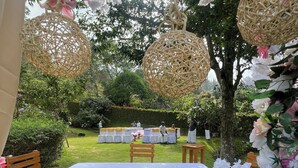 Un área de comedor al aire libre con una mesa blanca y sillas de madera, rodeada de exuberante follaje y lámparas decorativas de mimbre.