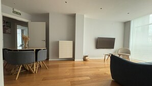 Una sala de estar moderna con piso de madera, un sofá, una mesa de comedor con sillas, un televisor montado en la pared y una puerta con la leyenda 'SALIDA'.