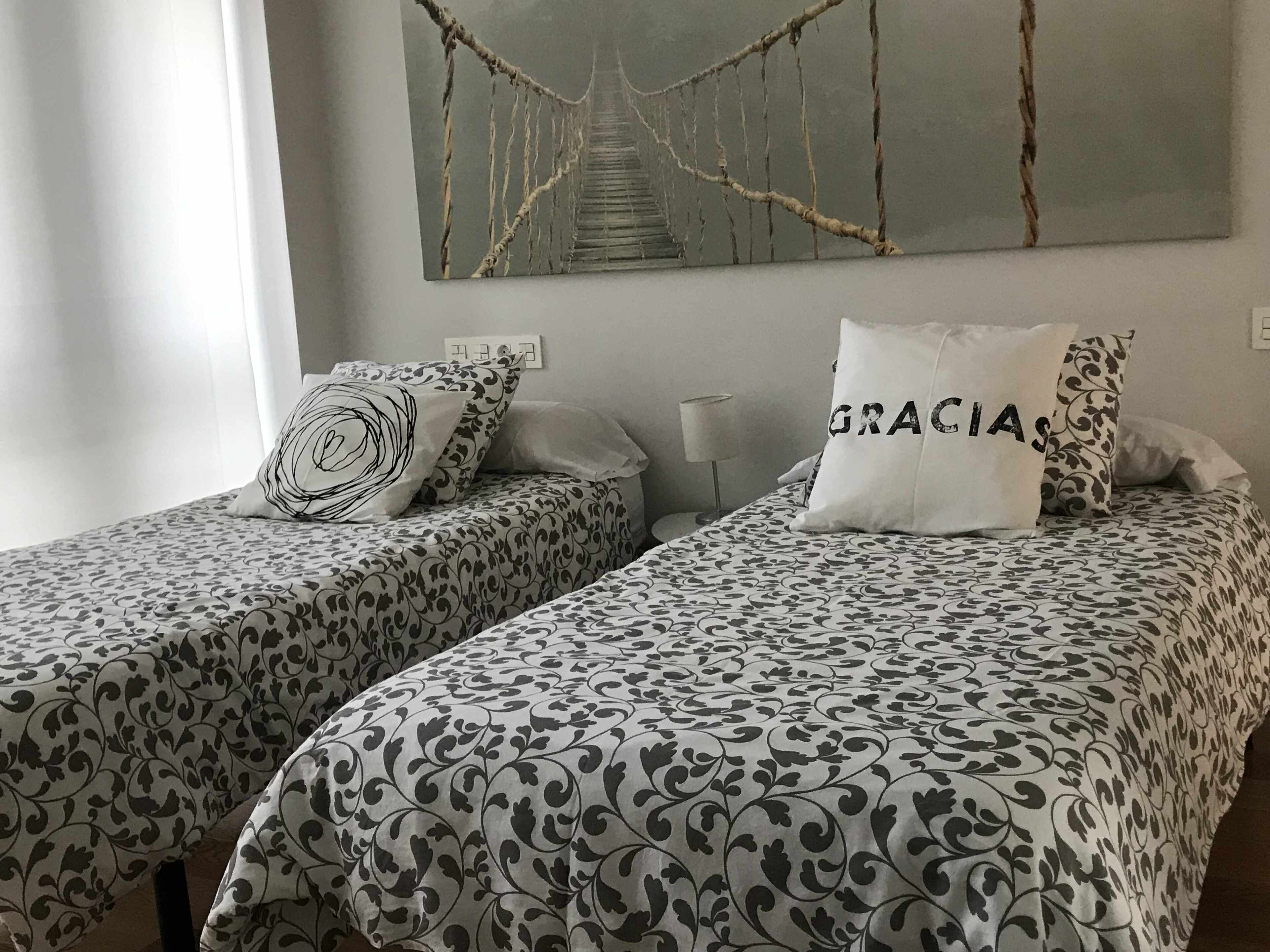 A hotel room with two beds, each with patterned bedding and decorative pillows. A painting of a bridge hangs on the wall above the beds.