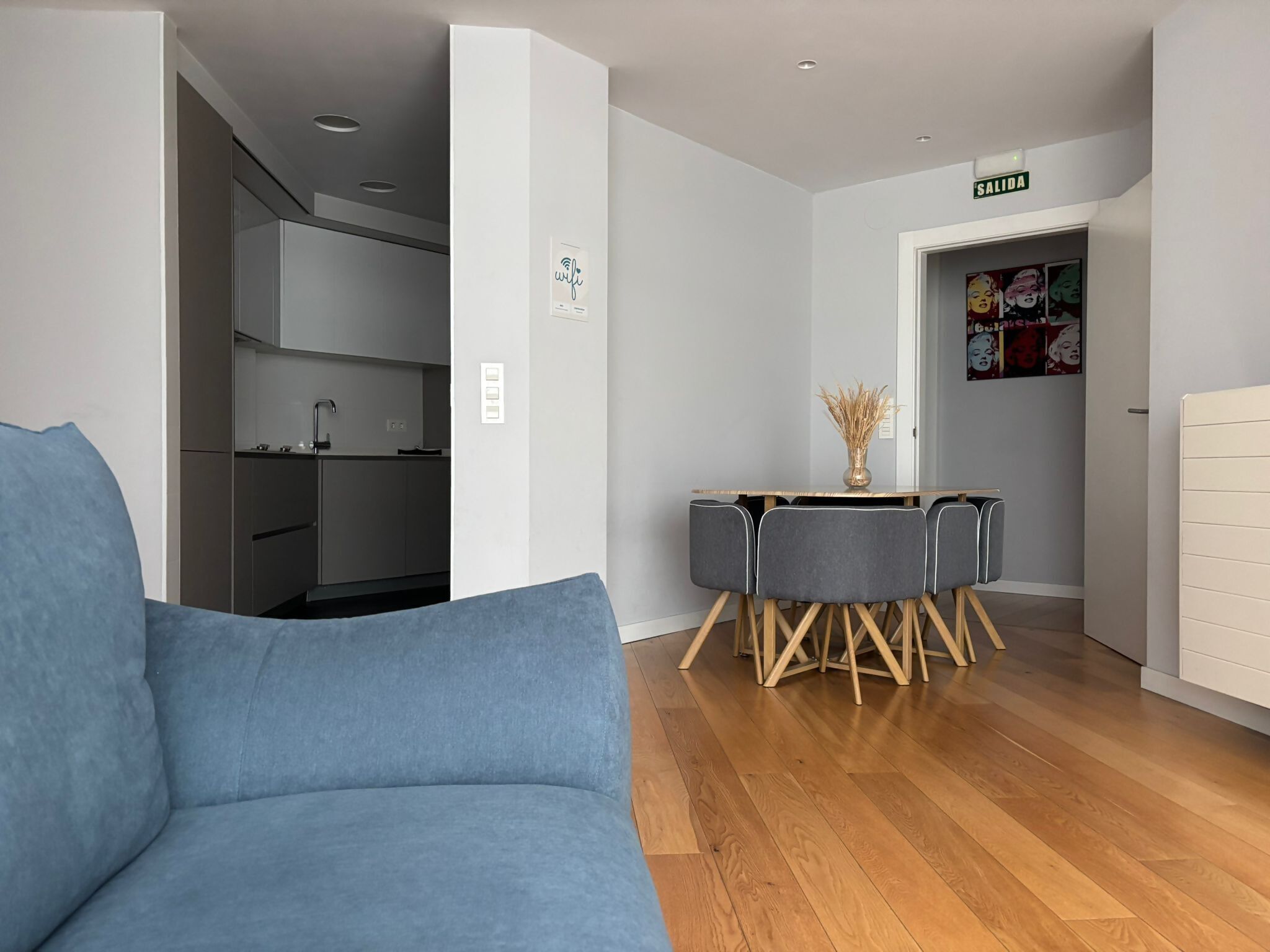 Una sala de estar moderna con un sofá azul, una mesa de comedor de madera con sillas, y una cocina al fondo.