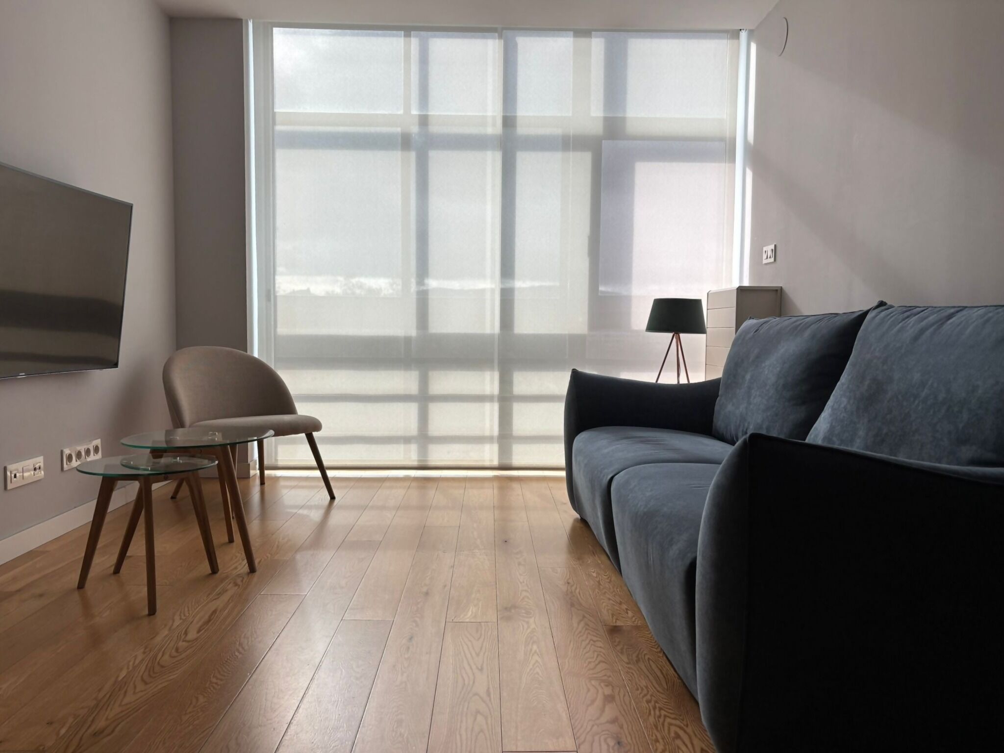 A modern living room with a large window, a flat-screen TV, a grey sofa, a small round table, and a lamp.