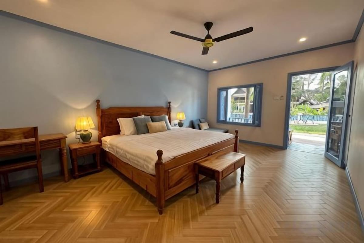 A bedroom with a wooden bed, bedside tables, a ceiling fan, and a view through a window.