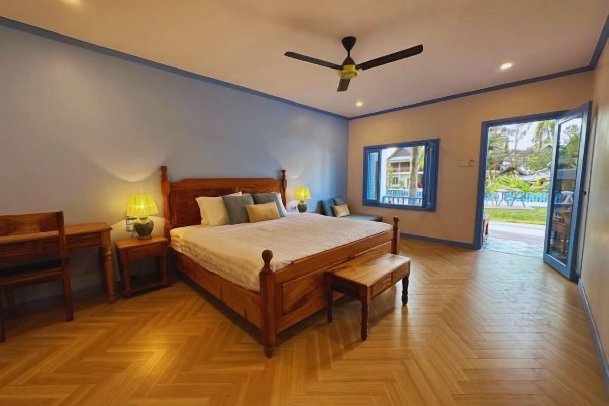 A bedroom with a wooden bed, a nightstand, a desk, a bench, a ceiling fan, and a large window.