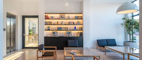A modern lobby with a glass coffee table, wooden chairs, a bookshelf, and a large window.