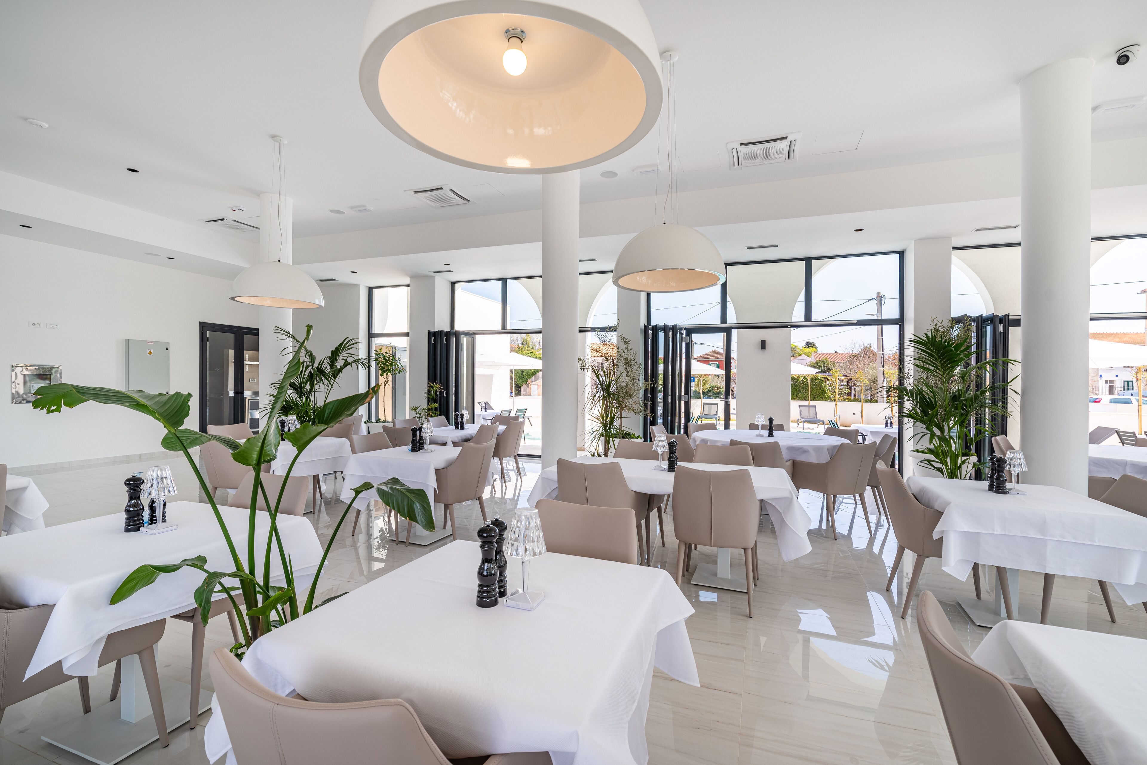 A spacious restaurant with high ceilings, large windows, and modern lighting fixtures. Tables are neatly arranged with white tablecloths and comfortable seating.
