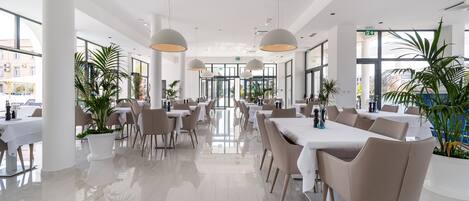 Un amplio comedor interior con varias mesas y sillas, ventanas grandes y plantas en macetas.