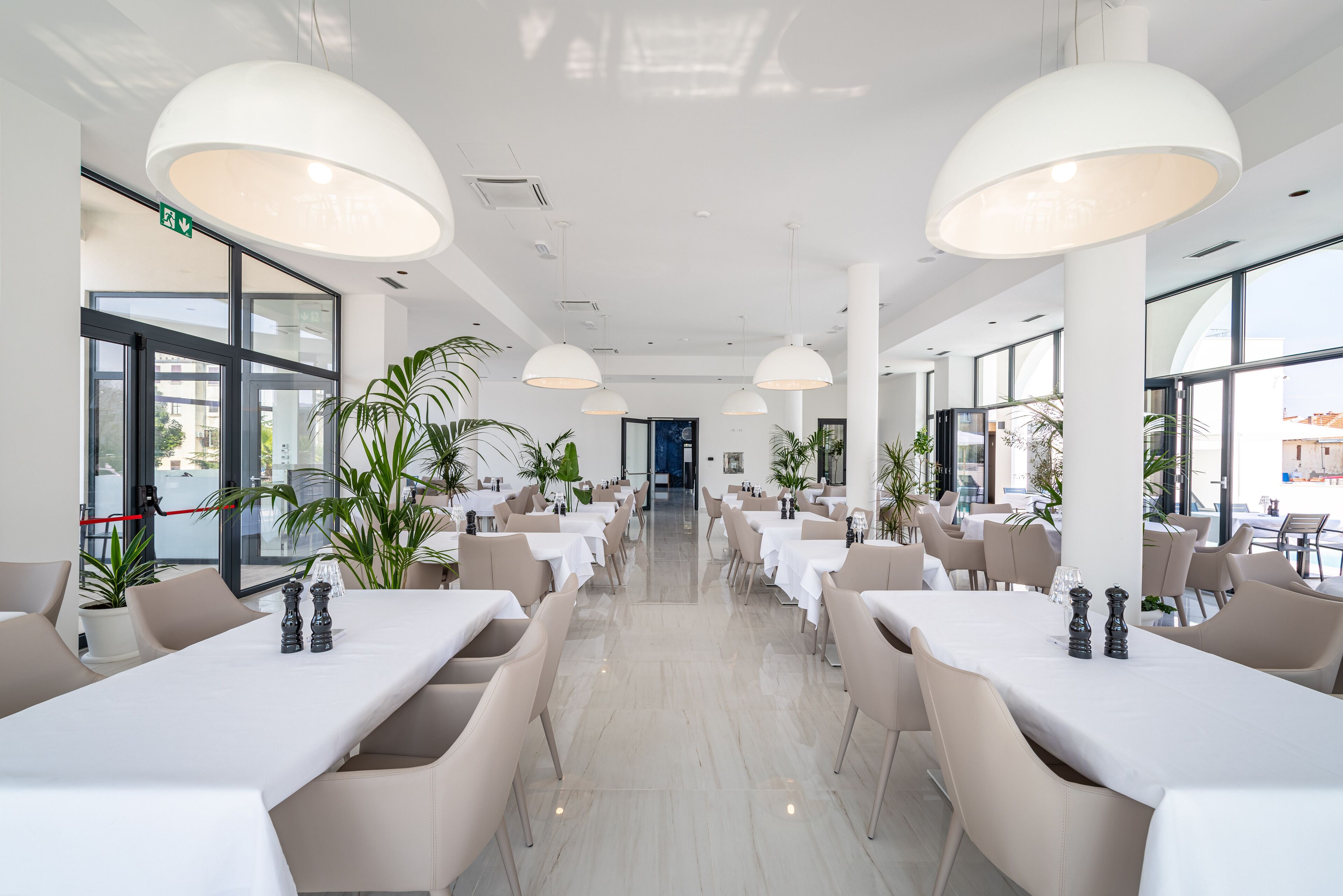 A spacious, modern dining area with high ceilings, large windows, and ample natural light. Tables are neatly arranged with white tablecloths and comfortable chairs.