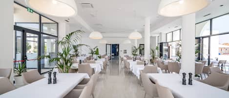 A spacious, modern dining area with high ceilings, large windows, and ample natural light. Tables are neatly arranged with white tablecloths and comfortable chairs.