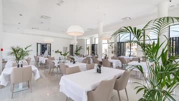 Un comedor amplio con varias mesas y sillas, ventanas grandes y plantas de interior.