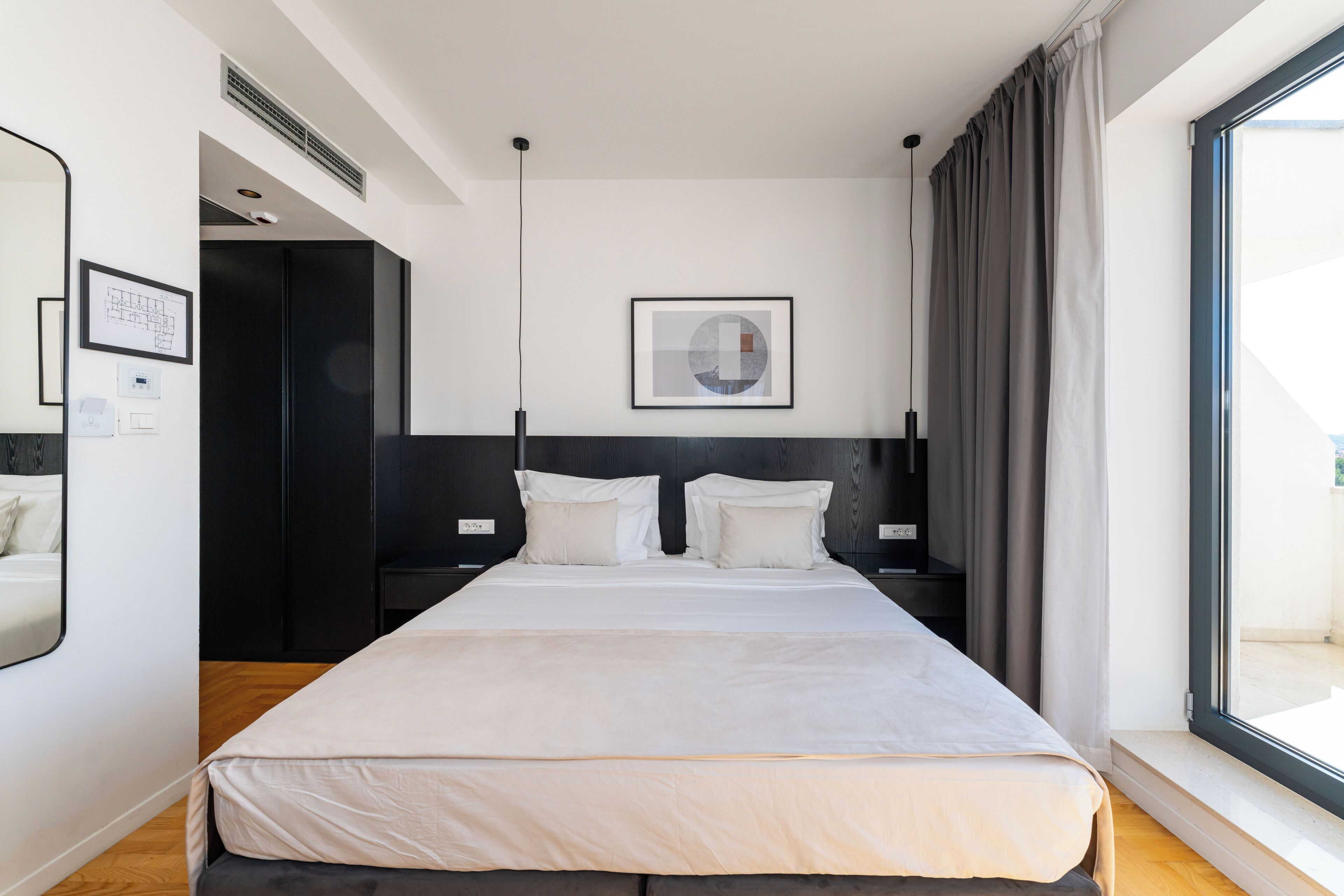 A modern hotel room with a large bed, a headboard, a bedside table, a nightstand, a window with curtains, and a wall-mounted artwork.